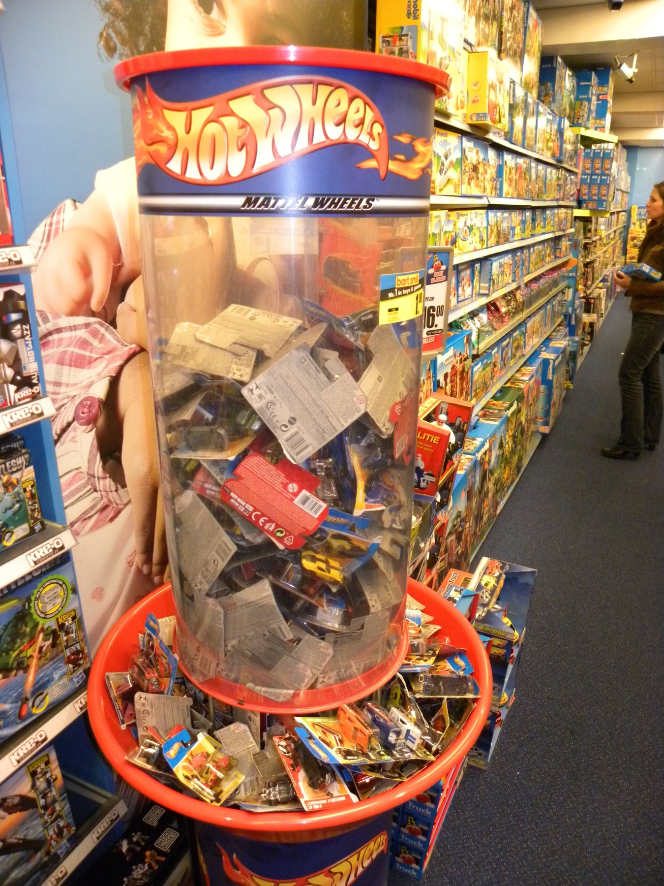 Hot-Wheels-Verkaufstonne voller verpackter Modellautos in einem Spielwarengeschäft, dahinter volle Regale