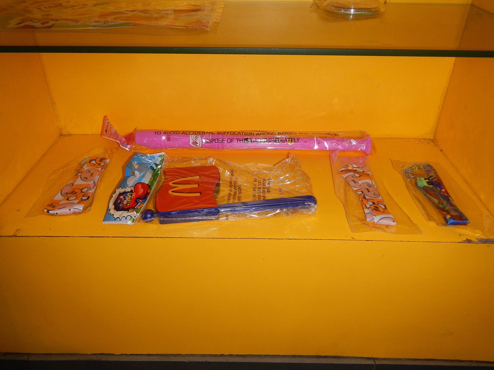 Mehrere Happy-Meal-Spielzeuge in ihren originalen Kunststoffbeuteln in einer gelben Vitrine